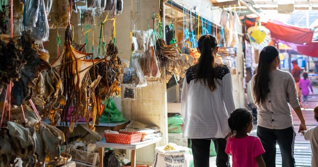 Women walking through a coastal market with various dried seafood products on display.