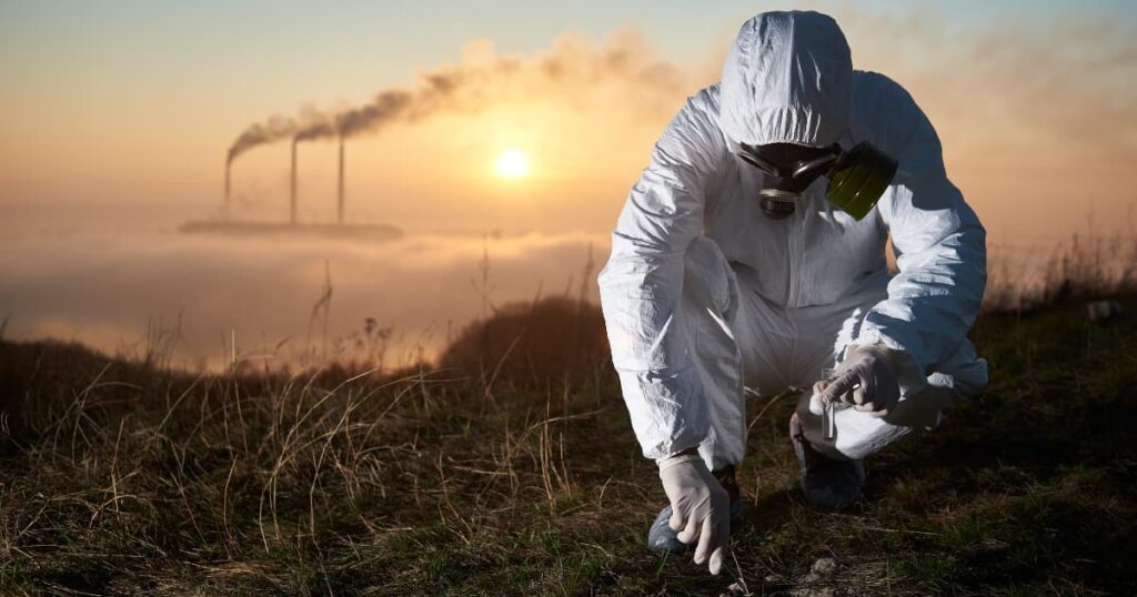 An environmental scientist in protective gear collecting soil samples near an industrial area.
