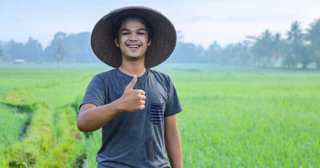 Young farmer smiling and giving a thumbs-up in a lush green field.