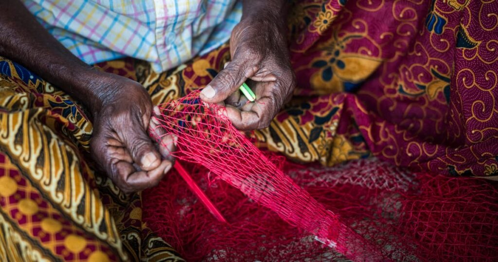 Close-up of elderly hands weaving a red net, showcasing traditional craftsmanship in coastal communities.